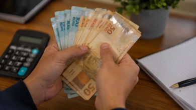 Pessoa contando notas de dinheiro em reais sobre uma mesa, com calculadora e caderno ao lado.