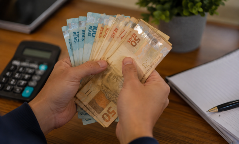 Pessoa contando notas de dinheiro em reais sobre uma mesa, com calculadora e caderno ao lado.